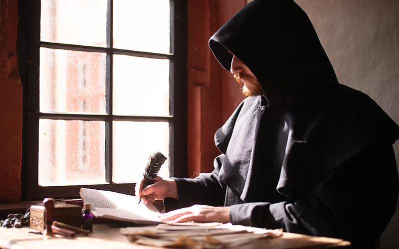 men sitting in a hooded cloak writing a letter with a feather