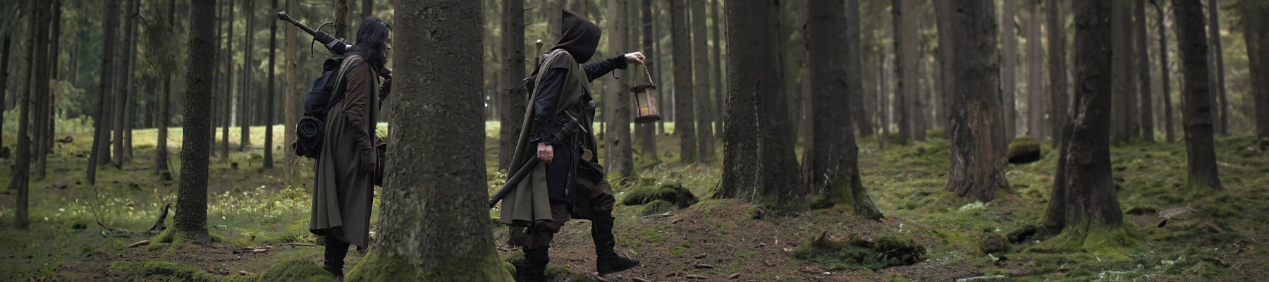 Two people dressed as rangers in dark green hooded clothing walking through a forest
