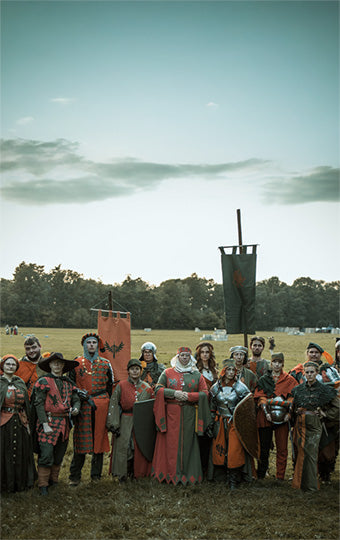 Group of people in medieval or fantasy costumes standing in a field with a banner in the background.