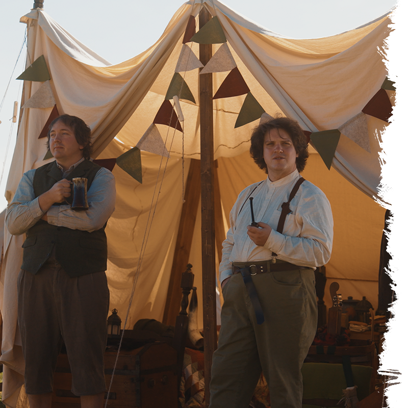 Two men in vintage attire standing in front of a large tent with decorative flags.