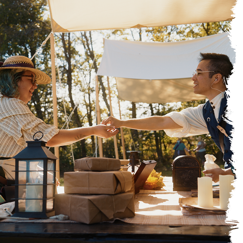 Two people shaking hands at a table with wrapped gifts and lanterns outdoors.