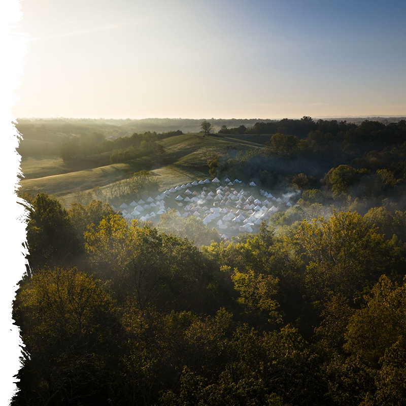 Aerial view of a field with tents surrounded by trees at sunrise or sunset.