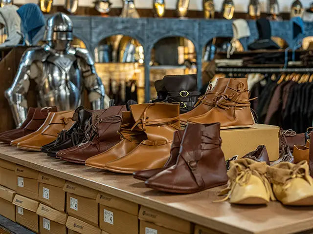 Row of brown medieval leather boots on a display table with armor and clothing in the background.