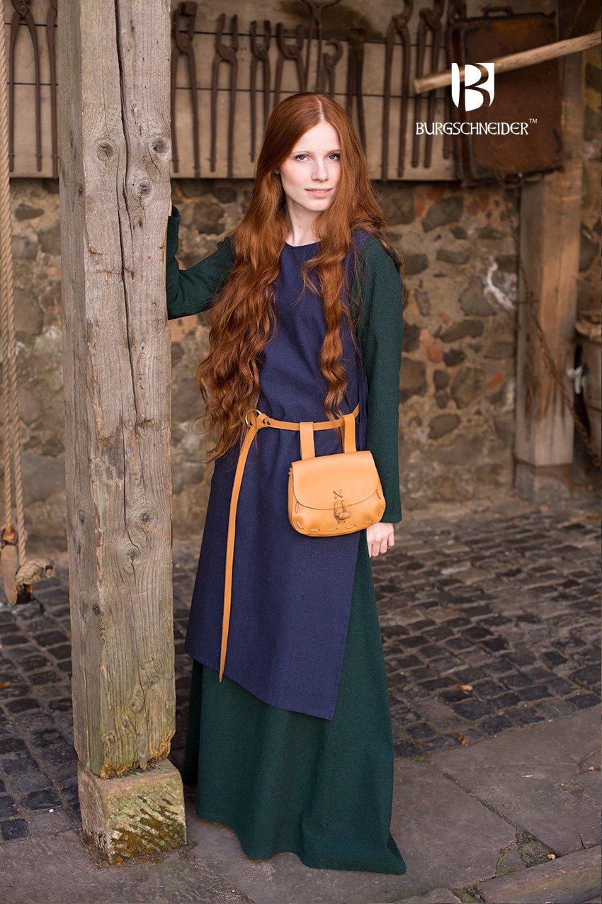 Woman in a medieval-style outfit with a blue dress and green cloak, standing against a stone wall.